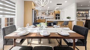 A modern dining area with a wooden table set for six, kitchen in the background, and a decorative chandelier.