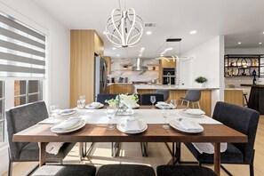 A modern dining area with a wooden table set for six, kitchen in the background, and a decorative chandelier.
