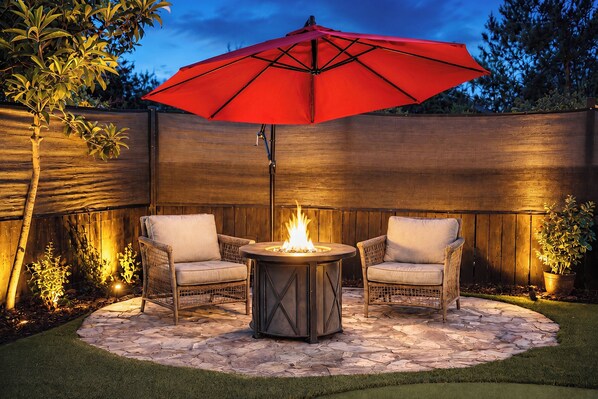 Une terrasse avec deux fauteuils en osier, une table ronde dotée d’un foyer et un parasol rouge.