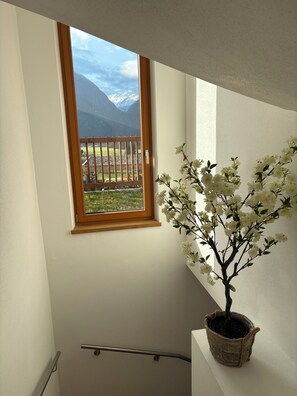 Eine Treppe mit einem hölzernen Geländer, ein Fenster mit Blick auf einen Holzzaun und Berge, und ein weiß blühender Topfpflanze.