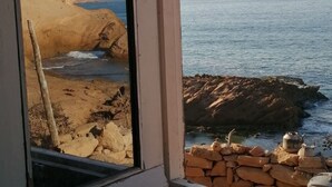 Uitzicht op de kust door een raam, met een zandstrand, rotsformaties en een heldere lucht.