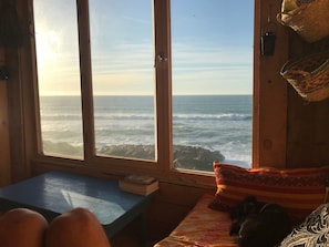 Une pièce dotée d’une grande fenêtre donnant sur l’océan, d’une table en bois et d’un lit agrémenté d’un coussin à motifs.