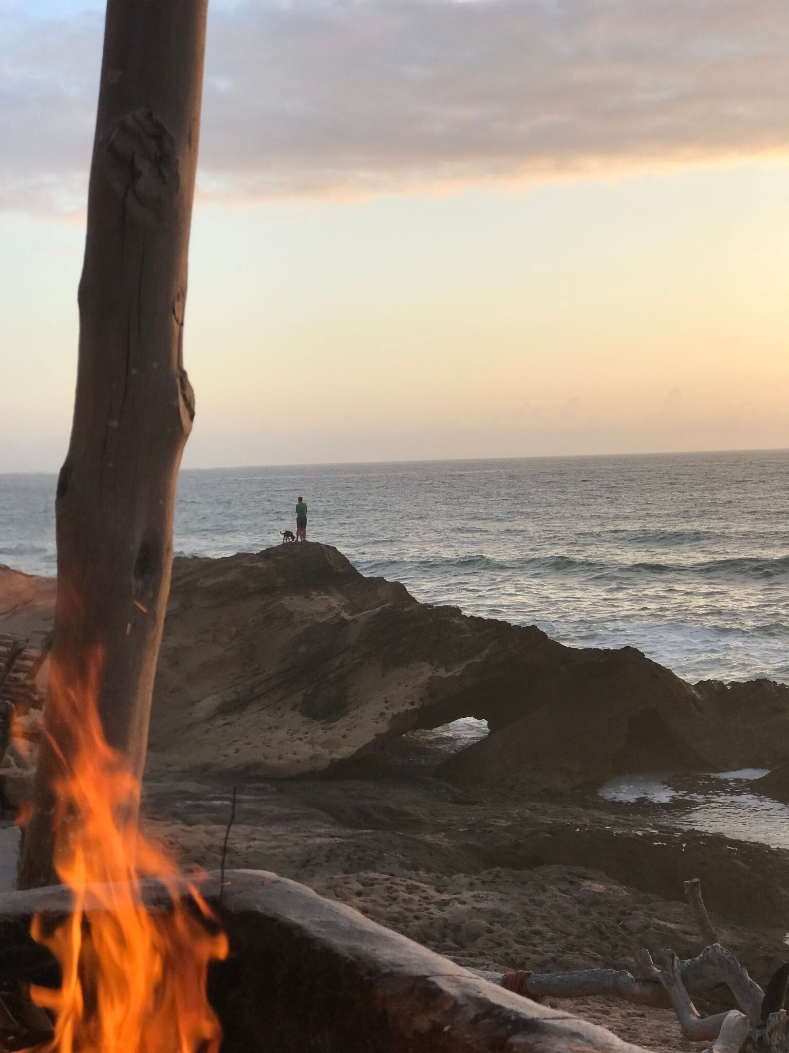 A beach scene with a fire, rocky terrain, and a person standing on a rock formation.