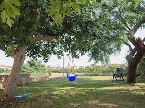 Ein Baum mit einer Schaukel, einem blauen Eimer und einem Liegestuhl auf einer Rasenfläche.
