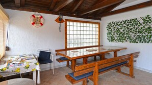 Ein Zimmer mit Holztisch, Bänken, einem fensterartigen Gitter und einer an der Wand befestigten Pflanze, sowie einer Rettungsboje als Dekoration.