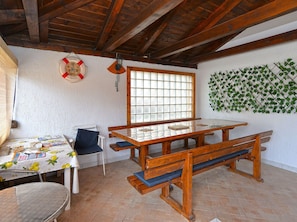 Ein Zimmer mit Holztisch, Bänken, einem fensterartigen Gitter und einer an der Wand befestigten Pflanze, sowie einer Rettungsboje als Dekoration.