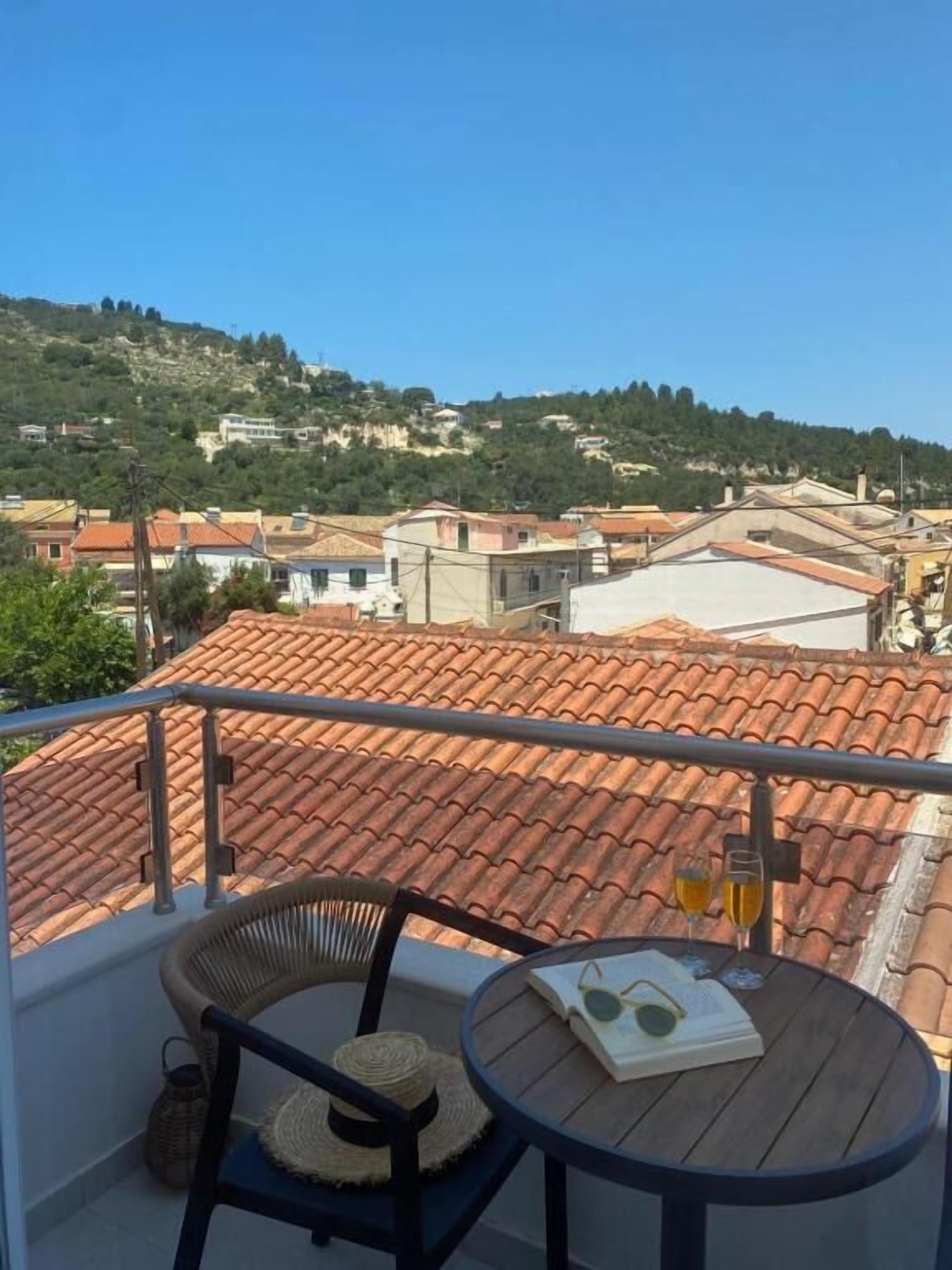 A balcony with a view of a town, a round table, a wicker chair, and a pair of sunglasses on the table.