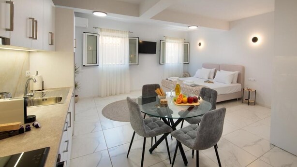 Quarto de hotel moderno com uma cama, uma mesa de jantar de vidro com cadeiras, uma cozinha e uma TV fixada na parede.