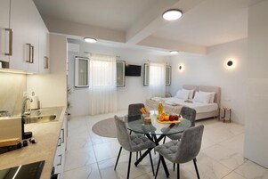 Una habitación de hotel moderna con una cama, una mesa de comedor de vidrio con sillas, una zona de cocina y un televisor montado en la pared.