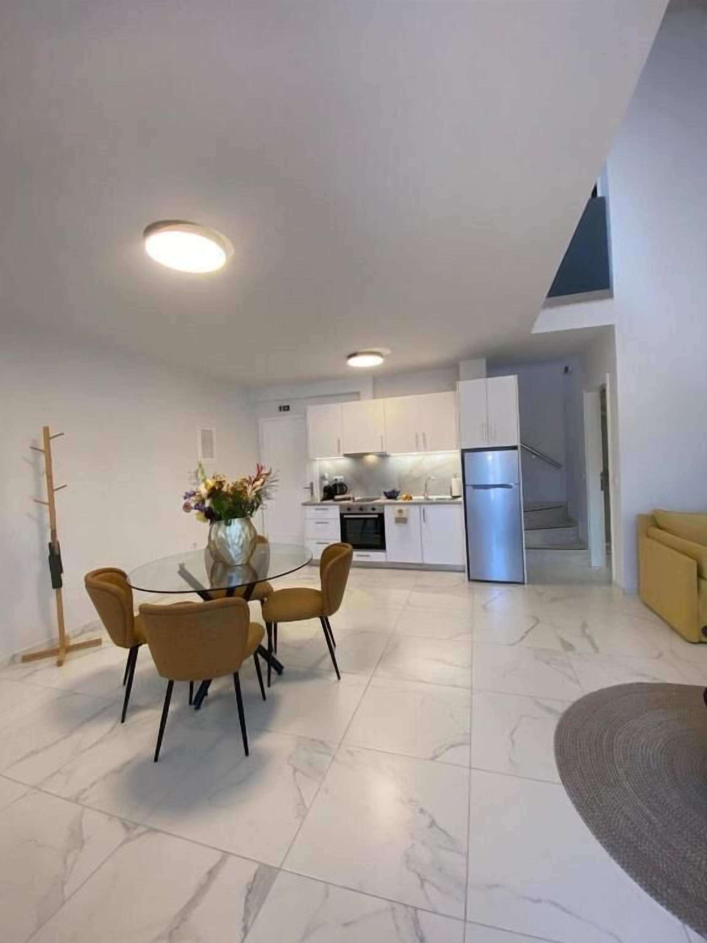 A modern kitchen with white cabinets, a stainless steel refrigerator, and a dining area with a glass table and four chairs.