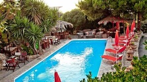 Una piscina rectangular con sombrillas rojas, rodeada de sillas y mesas de descanso, con vegetación y árboles al fondo.