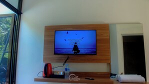 Ein an der Wand befestigter Fernseher mit einem Holzregal darunter, auf dem ein roter Lautsprecher, eine Wasserflasche und eine Fernbedienung stehen.