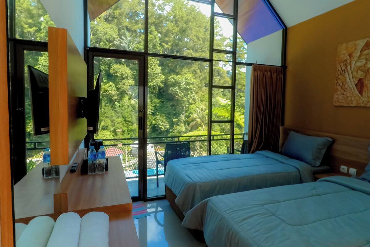 A bedroom with two beds, a desk with a flat-screen TV, and a large window with a view of trees.