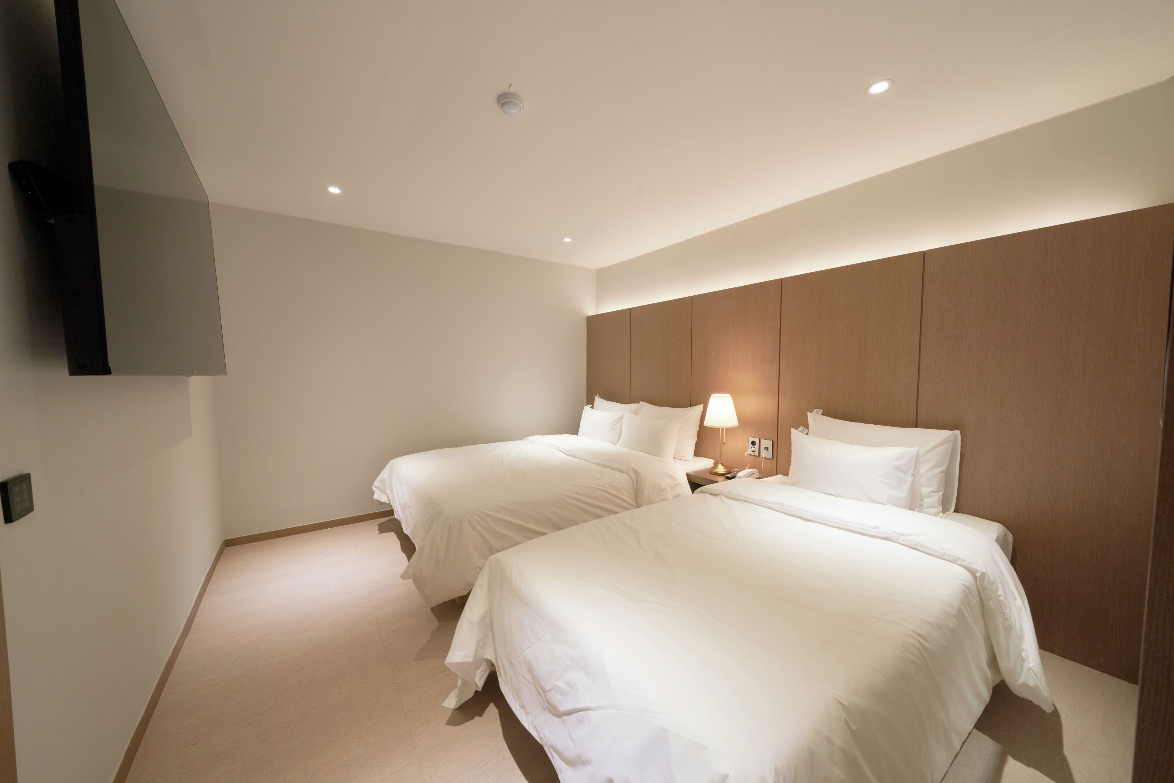 Habitación de hotel con dos camas, un televisor de pantalla plana montado en la pared y una mesita de noche con lámpara.