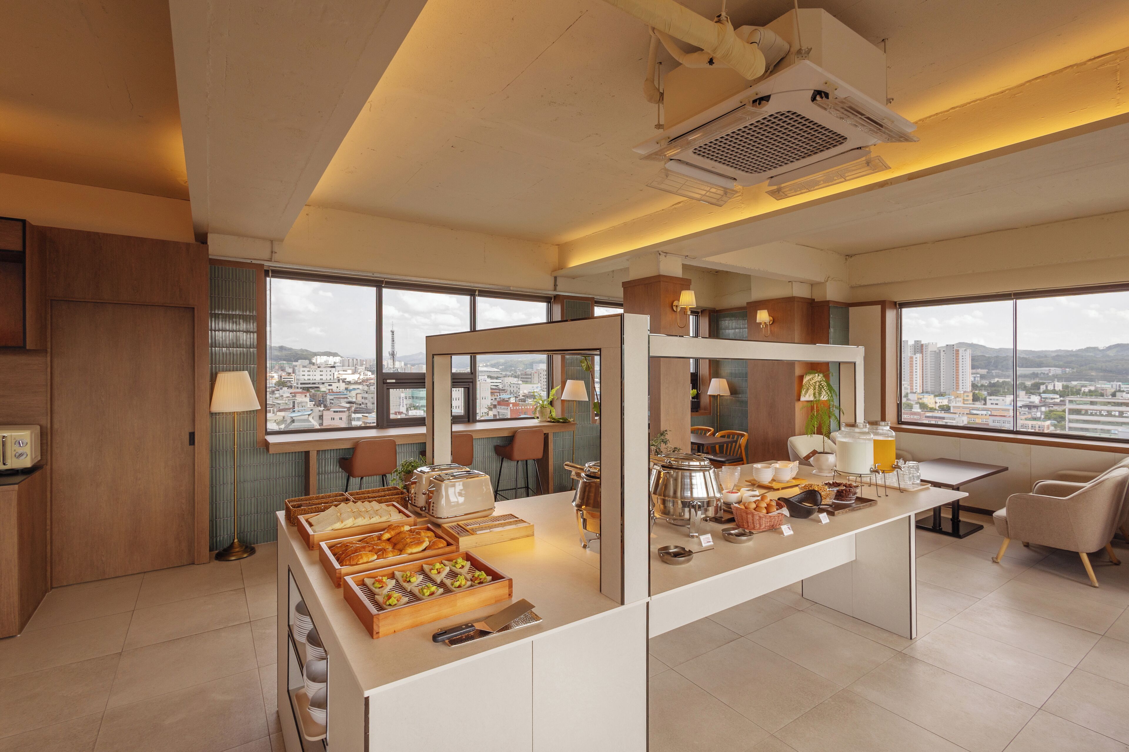 A modern hotel room with a breakfast bar, large windows with city views, and a kitchenette.