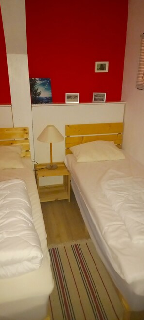 A hotel room with two single beds, a nightstand with a lamp, a red wall with framed pictures, and a striped rug.