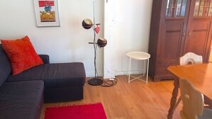 A living room with a dark sofa, red cushions, a framed picture on the wall, a floor lamp, a small white stool, and a wooden table. - Vacation apartment Glockenblume 5 (Siegsdorf)