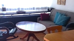 A dining area with a round table, wooden chairs, a sofa, and a window with blinds. - House Crocus, room no. 12 (Siegsdorf)
