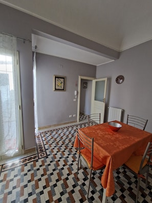 Ein Essbereich mit einem gemusterten Boden, einem Tisch mit roter Tischdecke, Metallstühlen, einer weißen Tür und einer Uhr an der Wand.