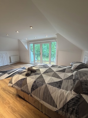 A bedroom with a large bed, a wooden floor, a view through a sliding glass door, and recessed ceiling lights.