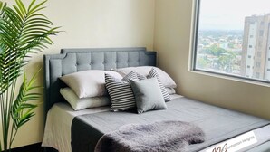Quarto com cabeceira estofada cinza, uma janela grande com vista para a cidade, uma planta em vaso e uma cama com roupa de cama cinza e almofadas estampadas.