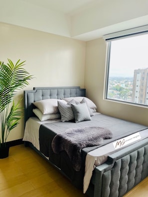 A bedroom with a grey tufted headboard, a large window with a city view, a potted plant, and a bed with grey bedding and patterned pillows. - Minimalist 1BR Condo in Araneta City (Quezon City)