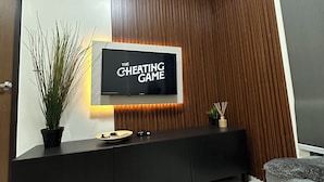 Uma sala de estar moderna com uma TV de tela plana exibindo 'The Cheating Game', um móvel de entretenimento de madeira escura, uma planta em vaso e um sofá cinza.