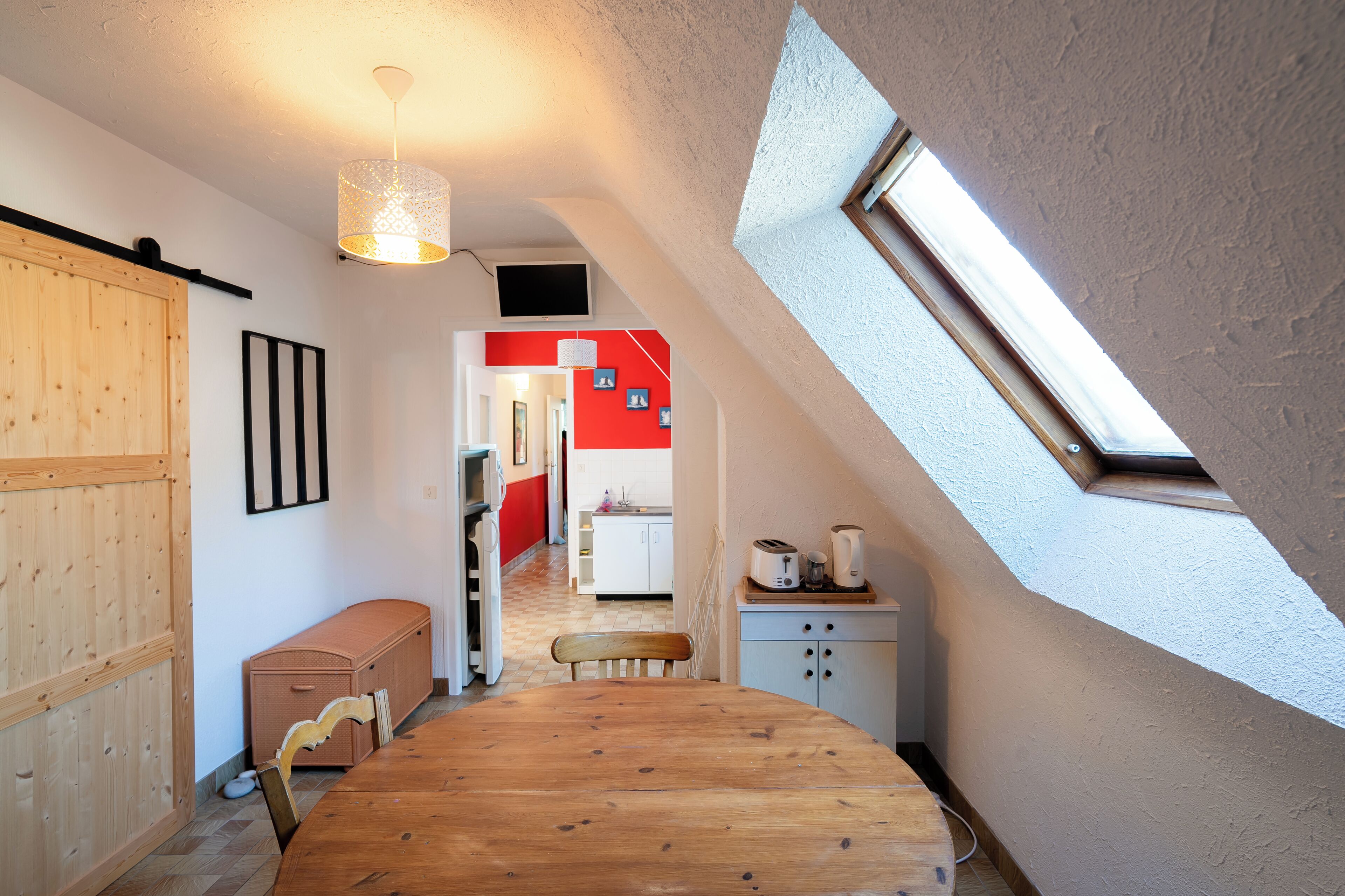 A room with a sloped ceiling, a wooden dining table, a bench, a small kitchenette, and a red accent wall.