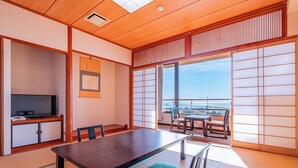 Ein traditioneller japanischer Raum mit Tatami-Böden, einem niedrigen Tisch, Stühlen, einem Fernseher und einer Schiebetür, die auf einen Balkon mit Blick führt.