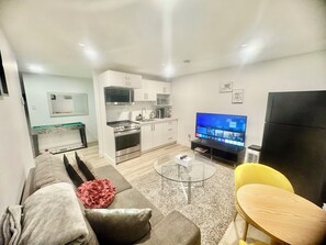 En moderne stue med sofa, sofabord, fjernsyn og et køkkenområde med skabe og hårde hvidevarer.