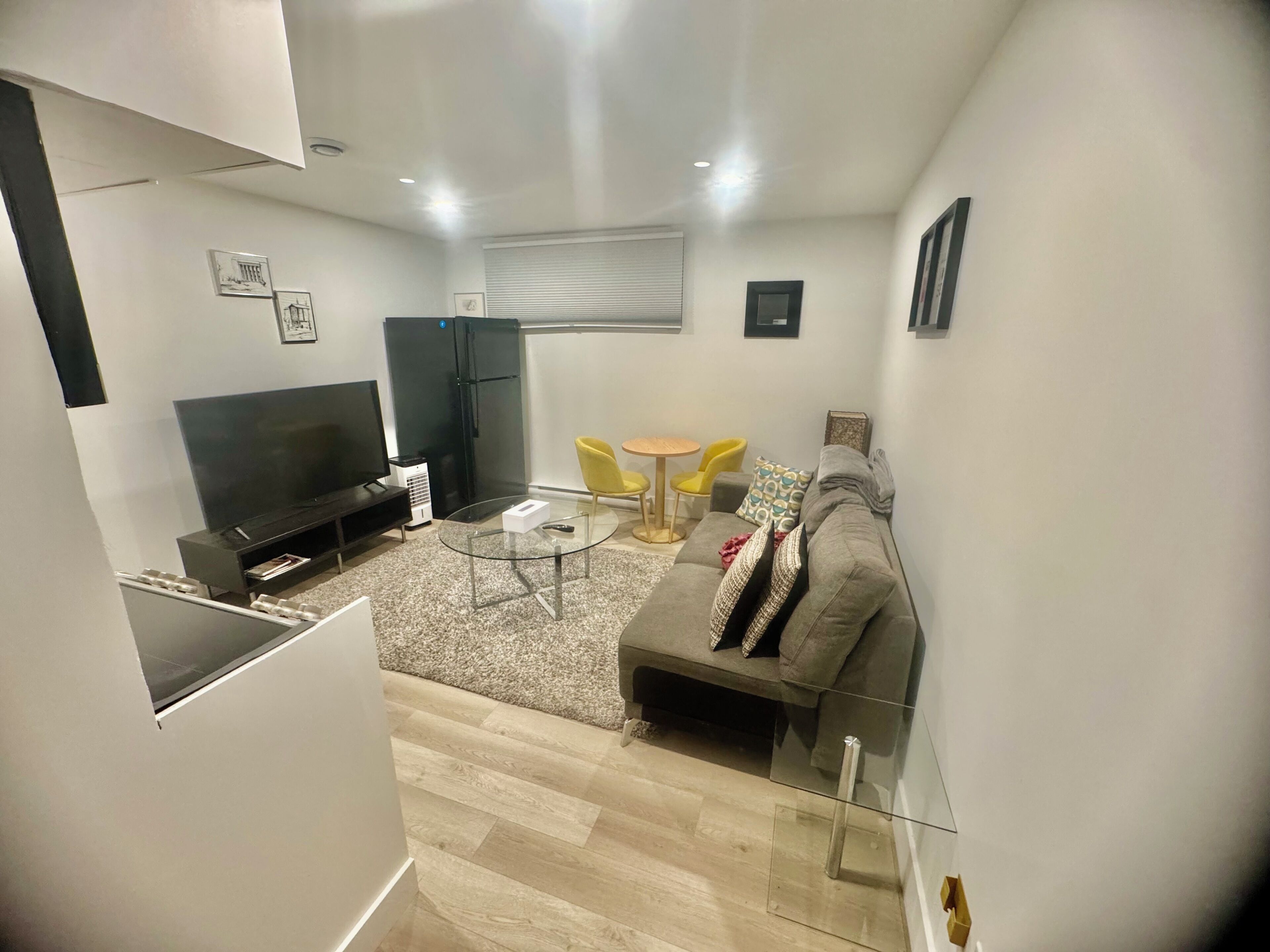 A modern living room with a flat-screen TV, a glass coffee table, a sofa, and a dining area with yellow chairs.