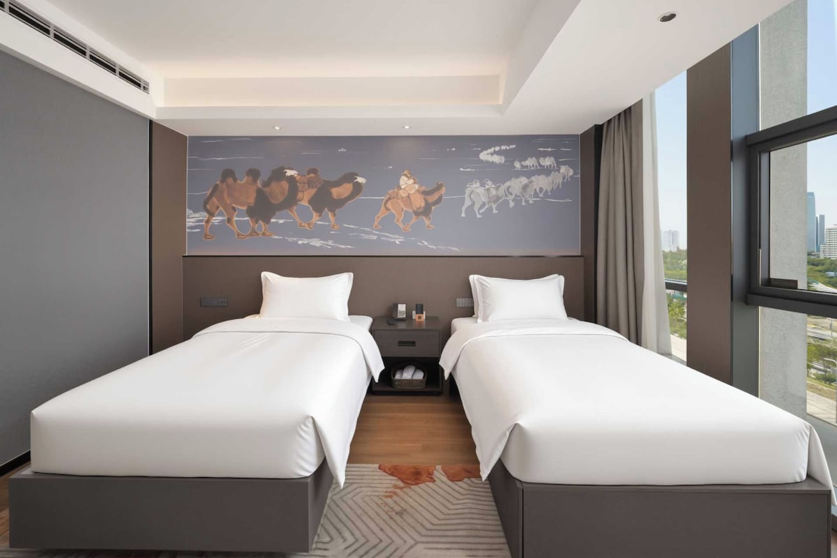 A hotel room with two beds, a large window, and a mural of camels on the wall.
