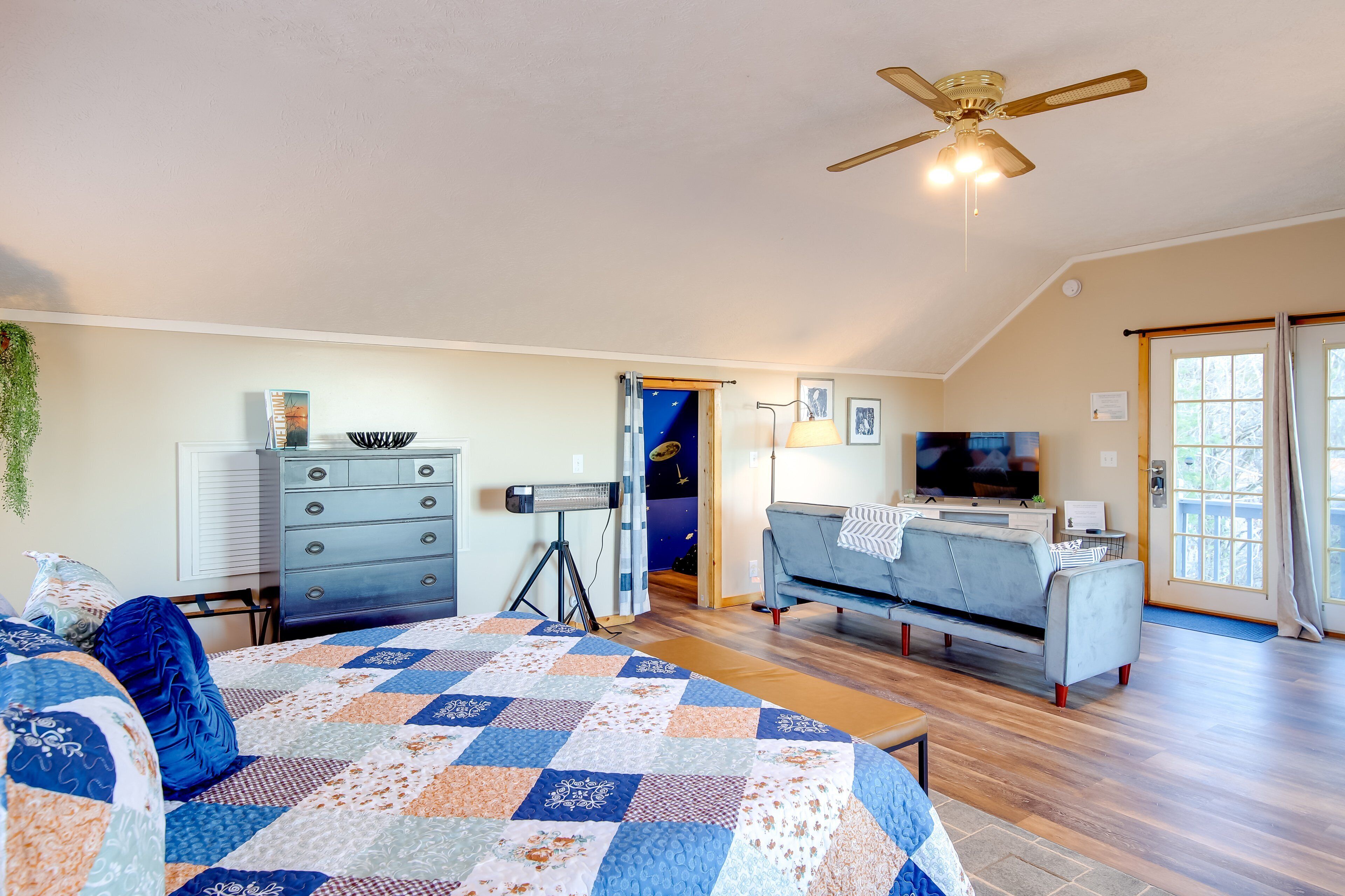 A bedroom with a bed, dresser, ceiling fan, and a TV in a living area with a couch and a window.