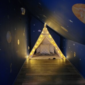 Una cama tipo tienda de campaña con decoraciones de estrellas y luces colgantes, frente a una pared azul oscuro con motivos celestes.