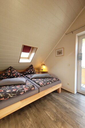 Ein Schlafzimmer im Loft-Stil mit schrägem Dach, einem Oberlicht, einem Bett mit floralem Bettbezug, einem Nachttisch mit Lampe und einer Tür, die nach draußen führt.