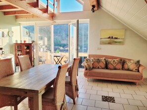 Zona pranzo con tavolo in legno e sedie in vimini, divano con cuscini decorati e vista su un patio esterno attraverso ampie vetrate.