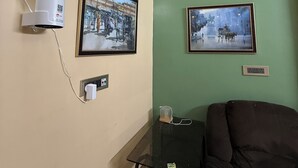 A room with a beige wall, a green accent wall, a black table with a glass top, a black sofa, a white air conditioning unit, and two framed pictures. - The Ganapati Abode (Kolkata)