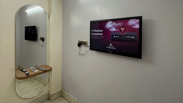 A bathroom with a mirror, a wall-mounted TV displaying a weather forecast, and a wooden shelf with personal items. - The Ganapati Abode (Kolkata)