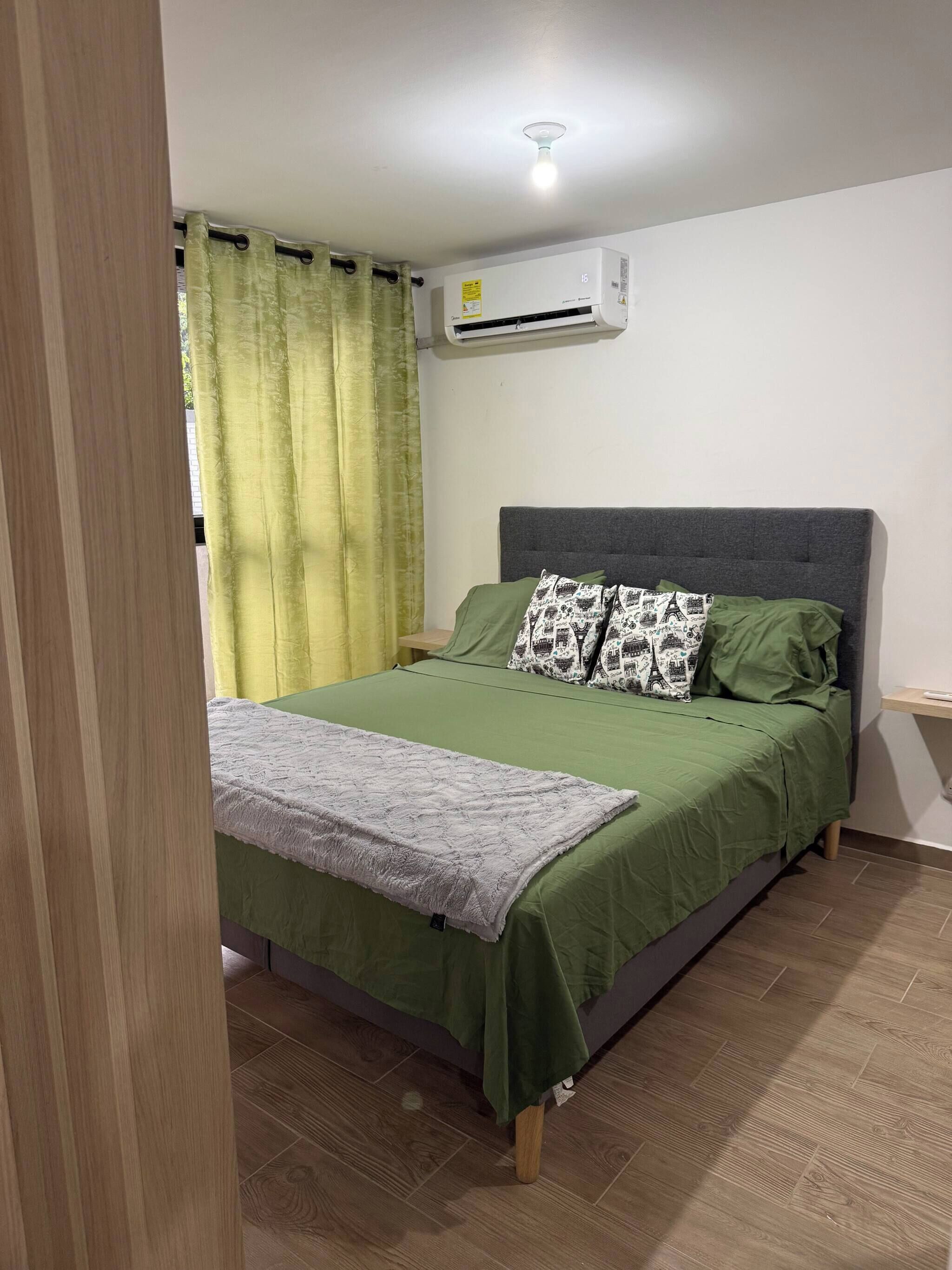 Ein Schlafzimmer mit einem Bett, grüner Bettwäsche, einem grauen Kopfteil, einer grauen Decke, einem Kopfkissen mit Stadtansicht-Motiv, einem grünen Vorhang und einer Klimaanlage an der Wand.