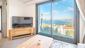 En moderne stue med et stort fjernsyn på et træstel, et sofabord og en balkon med havudsigt.