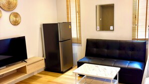 A modern living room with a flat-screen TV, a black refrigerator, a black sofa, a white coffee table, wooden flooring, and an air conditioning unit.