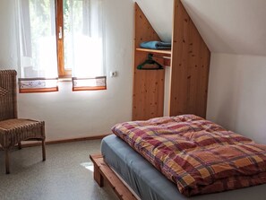 Ein Schlafzimmer mit einem Bett, einem Sessel, einem Holzregal und einem Fenster mit Vorhängen.