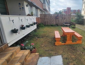 En privat trädgård med ett träbord och bänkar, en vit vägg med upphängda blomkrukor och ett bambustaket.