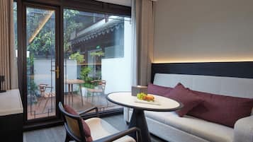 Quarto de hotel moderno com sofá, mesa redonda com uma cesta de frutas, cadeira e vista através de uma porta de vidro para uma área externa com plantas e um edifício tradicional.