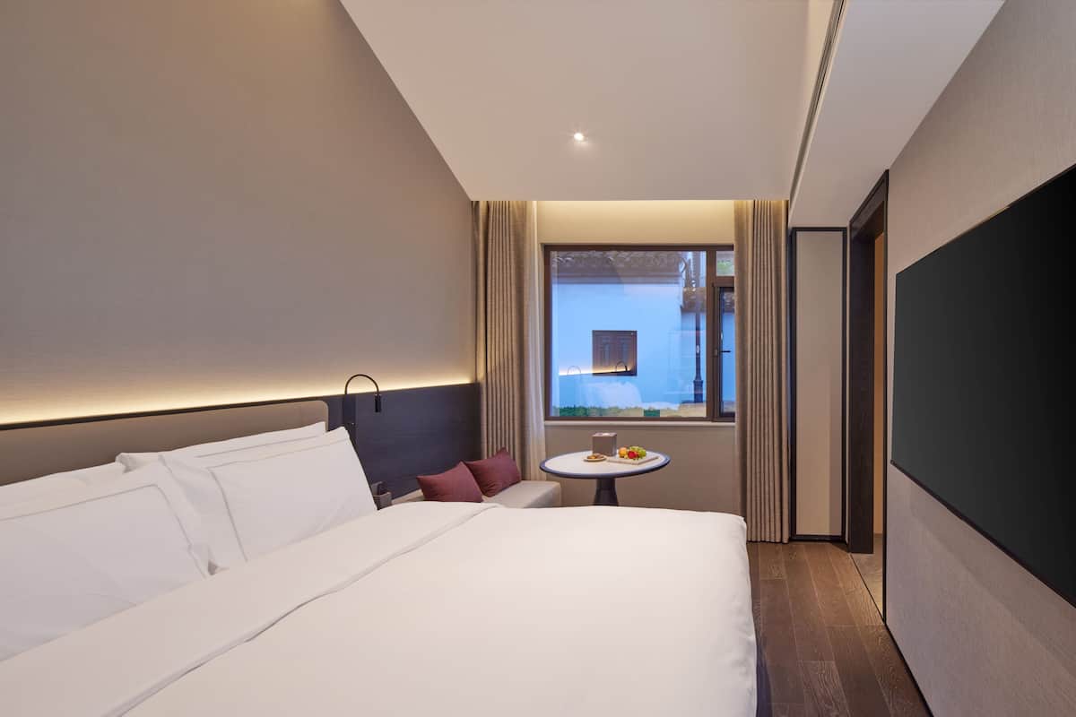 Ein Hotelzimmer mit einem großen Bett, einem kleinen runden Tisch mit Obstteller, einem Fenster mit Vorhängen und Blick auf ein Gebäude.