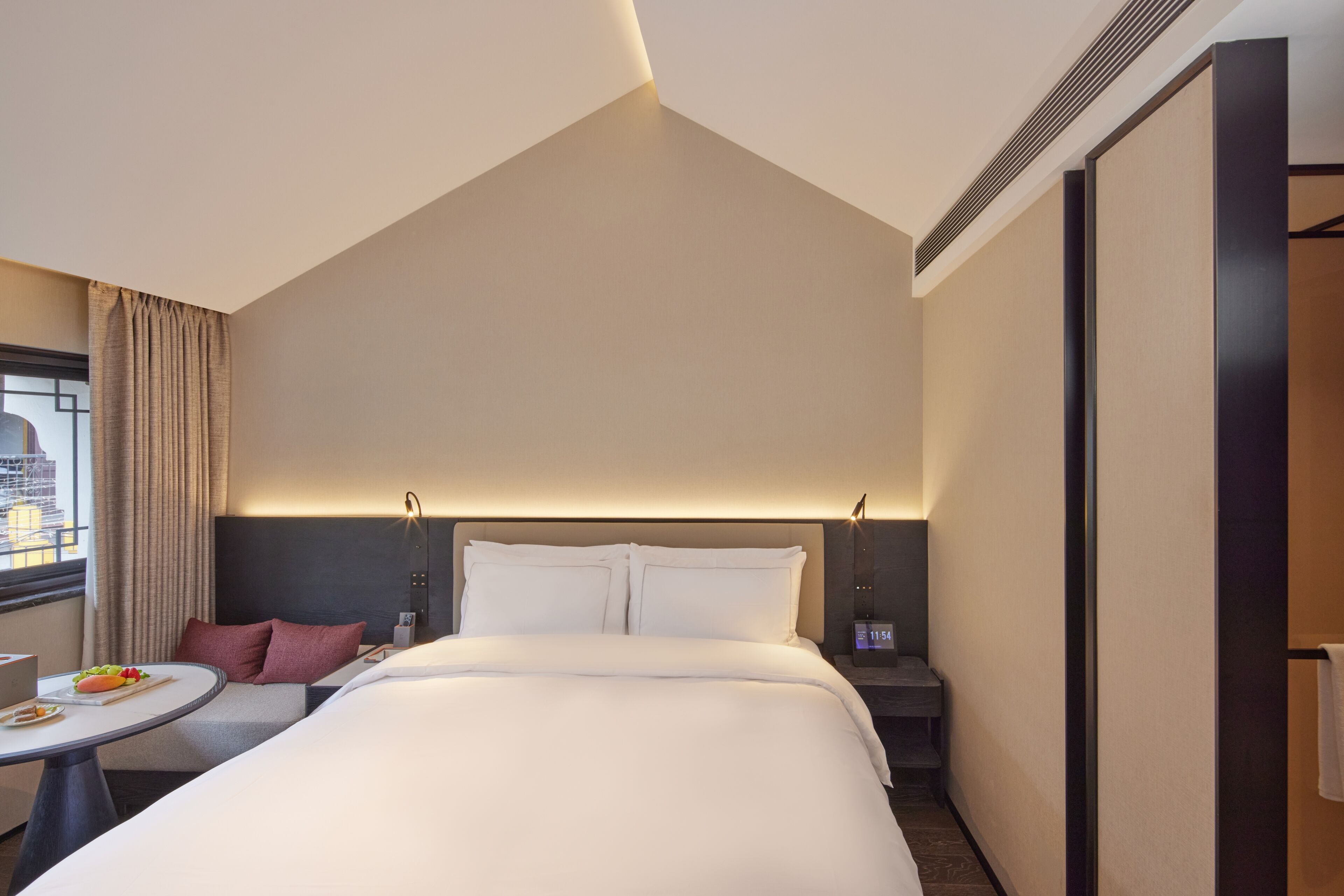 A modern hotel room with a large bed, bedside tables, a small round table with a fruit bowl, and a view of the city through a window.