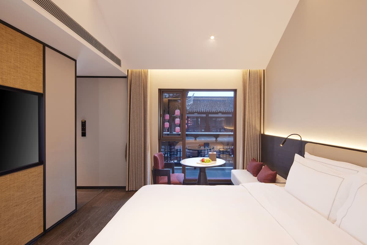 Ein Hotelzimmer mit Bett, Fernseher, Esstisch mit Obstschale und Blick auf ein traditionelles Gebäude durch ein Fenster.