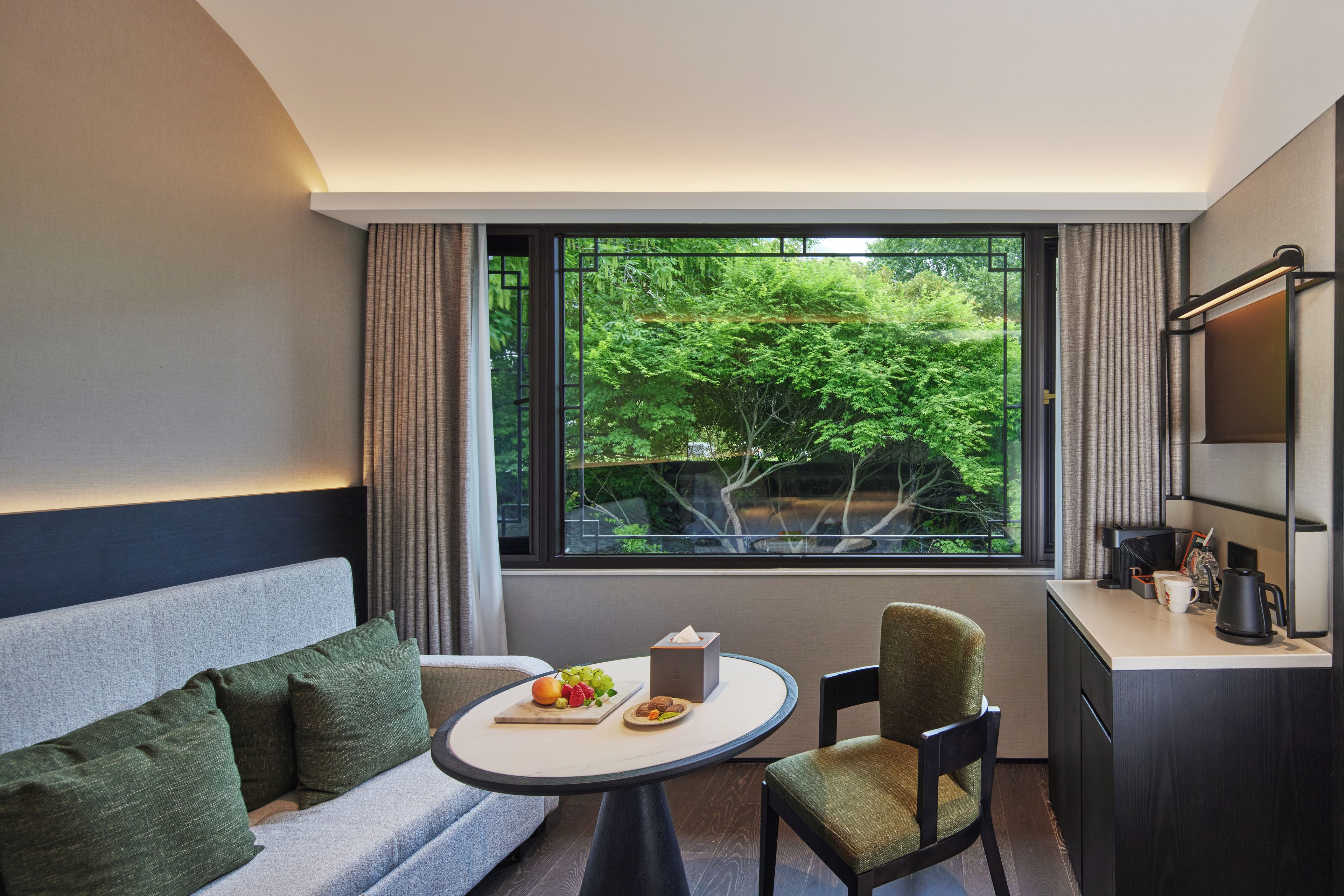 Une chambre d’hôtel moderne avec un canapé, une petite table ronde sur laquelle sont disposés des fruits, une chaise, un bureau avec une lampe et une grande fenêtre donnant sur un paysage verdoyant.