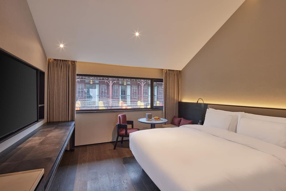 Ein Hotelzimmer mit einem großen Bett, einem Schreibtisch mit Stuhl, einem kleinen Tisch und einem Fenster mit Vorhängen.