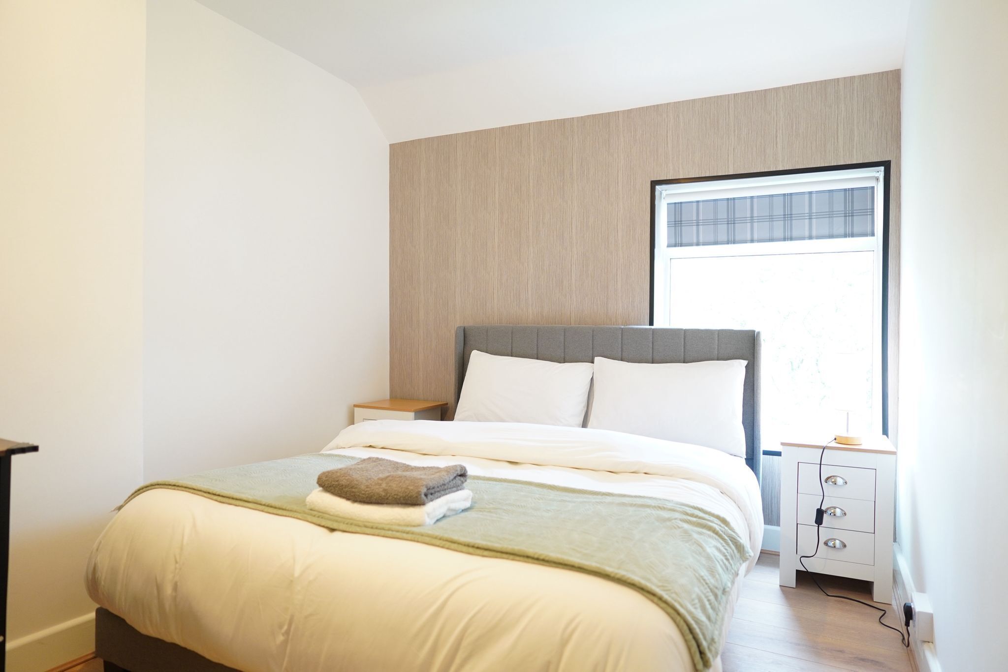 A neatly made bed with a grey headboard, a bedside table with drawers, and a window with blinds.